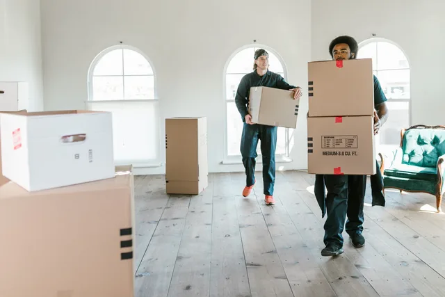 Two movers carrying boxes in a sunlit room, ready to assist with relocation. Flytt och transport: avtal, ansvar och krav